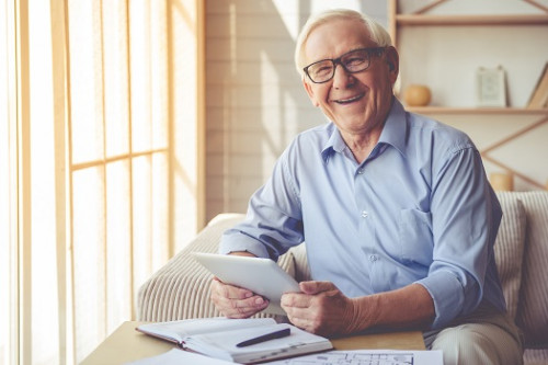 Handsome old man in eyeglasses is using a digital tablet, looking at camera and smiling while workin